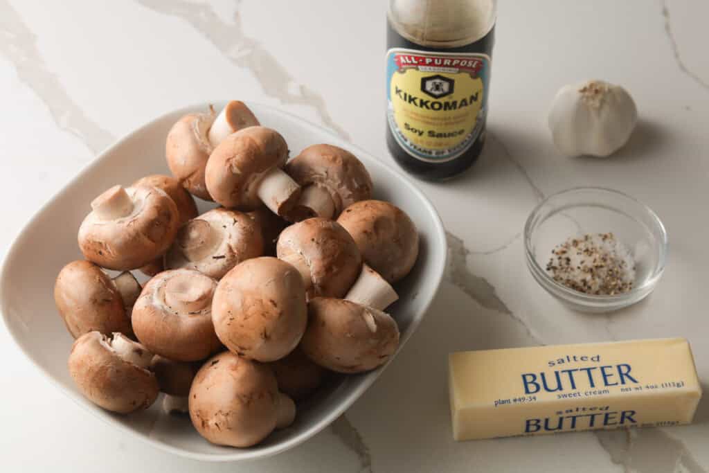 Ingredients gathered on a counter: a bowl of whole brown mushrooms, a stick of butter, a bottle of soy sauce, a bulb of garlic, and a small dish of salt and pepper.