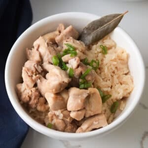 A square-cropped overhead shot of a white bowl filled with cooked chicken adobo and rice, garnished with fresh chopped green onions and a whole bay leaf.