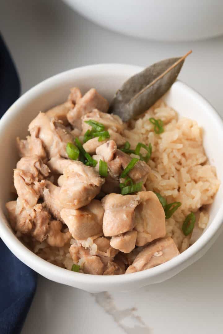 A vertical shot of chicken adobo and seasoned rice served in a white bowl, topped with sliced green onions and a bay leaf.