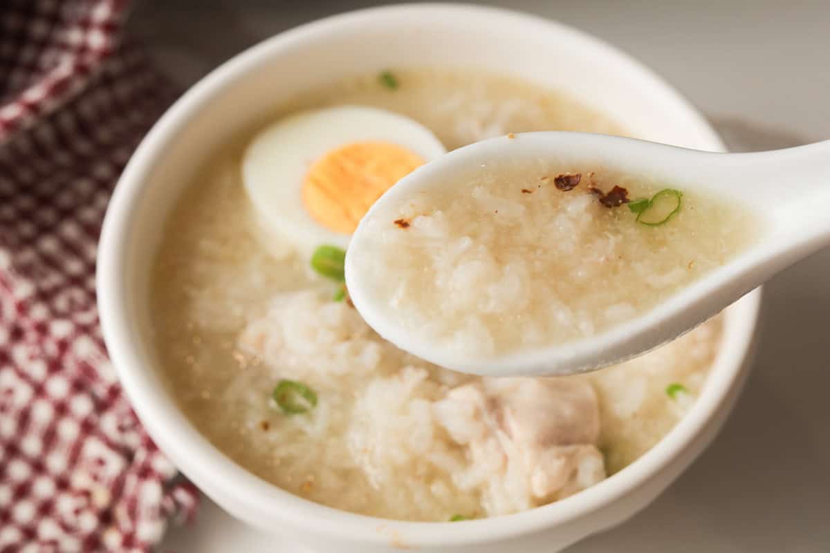 A white ceramic spoon scooping a portion of glistening chicken rice porridge from a bowl topped with a hard-boiled egg.