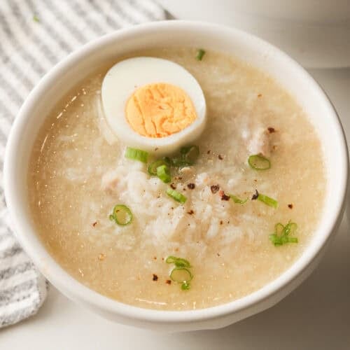 A close-up, top-down view of a white bowl filled with arroz caldo chicken rice porridge, topped with a hard-boiled egg half, sliced green onions, and black pepper.