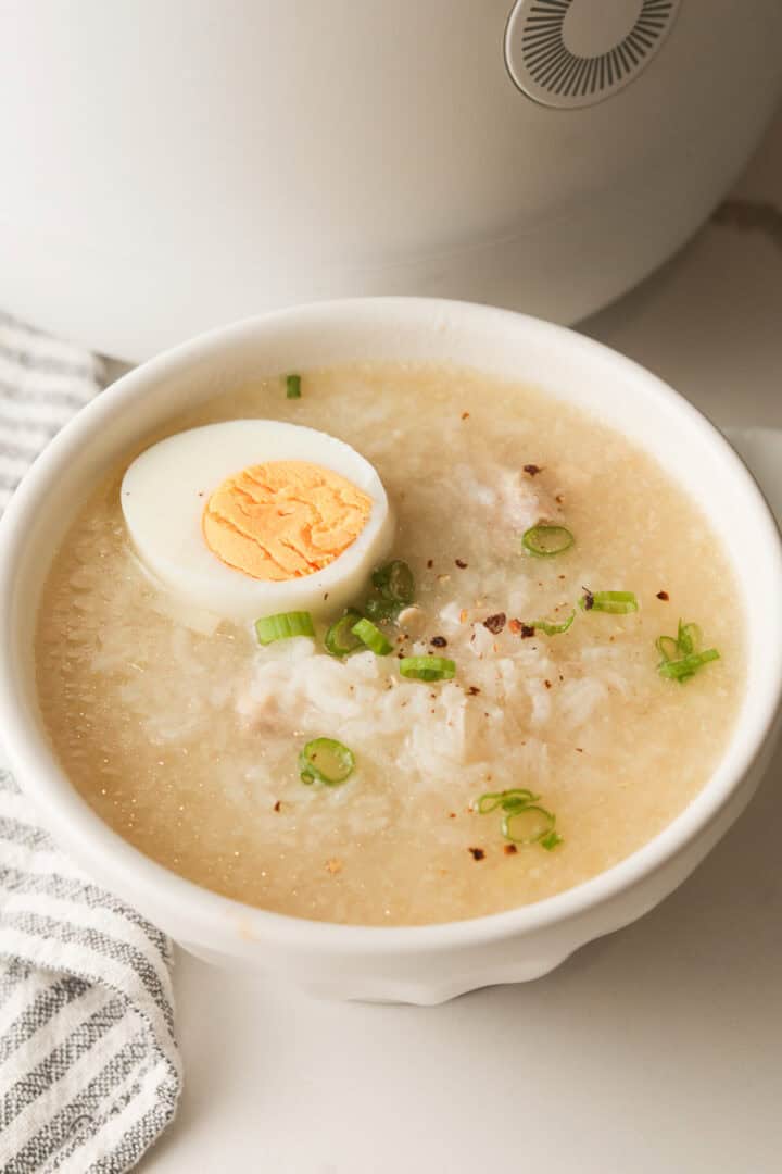 A bowl of chicken rice porridge with a hard-boiled egg and scallions, set against the blurred background of a white rice cooker.