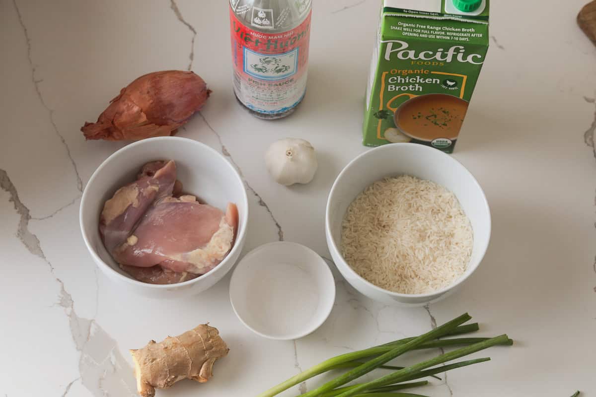 Raw ingredients arranged on a white countertop: bowls of raw chicken thighs and white rice, a carton of chicken broth, a bottle of fish sauce, an onion, a bulb of garlic, ginger root, and green onions.