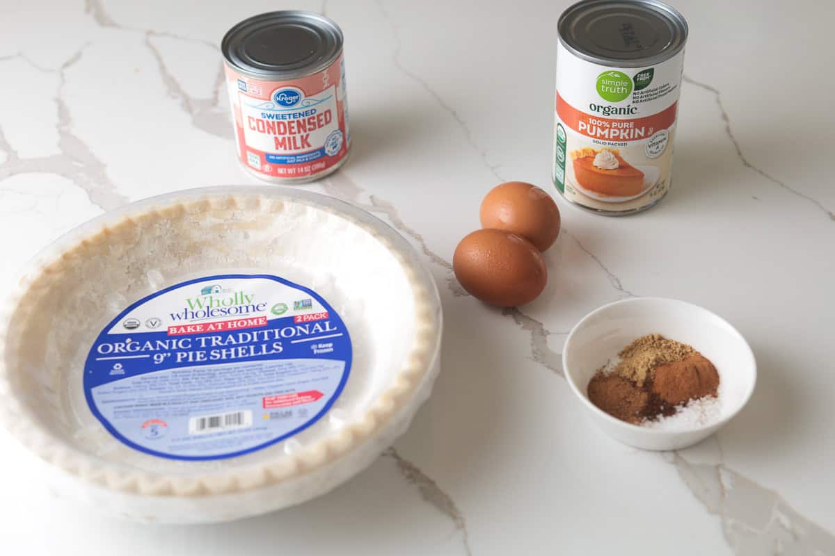 Canned pumpkin, sweetened condensed milk, eggs, spices, and a pre-made pie crust on a counter.
