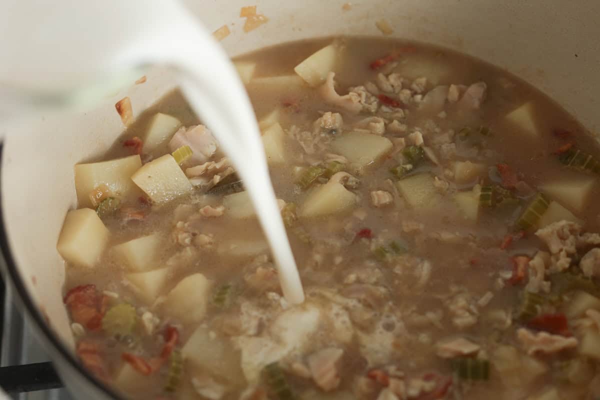 Heavy cream pouring into a pot of simmering clam chowder broth and potatoes.