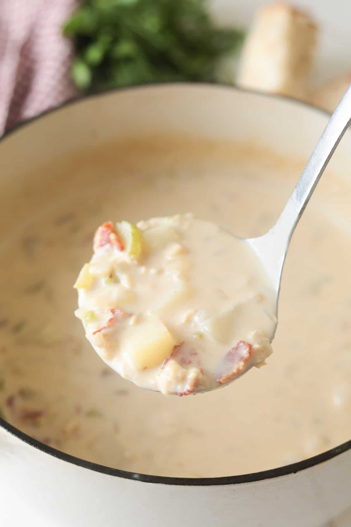 Metal ladle scooping up creamy clam chowder filled with potatoes, clams, and bacon.