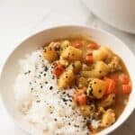 A close-up white bowl filled with white rice and hearty chicken curry containing chunks of potatoes and carrots, topped with black sesame seasoning, against a light marble background.