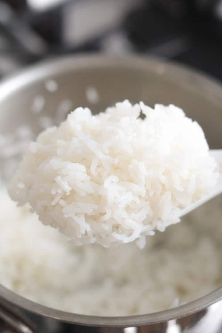 Vertical close-up shot of fluffy cooked jasmine rice on a rice paddle held over a stainless steel cooking pot.