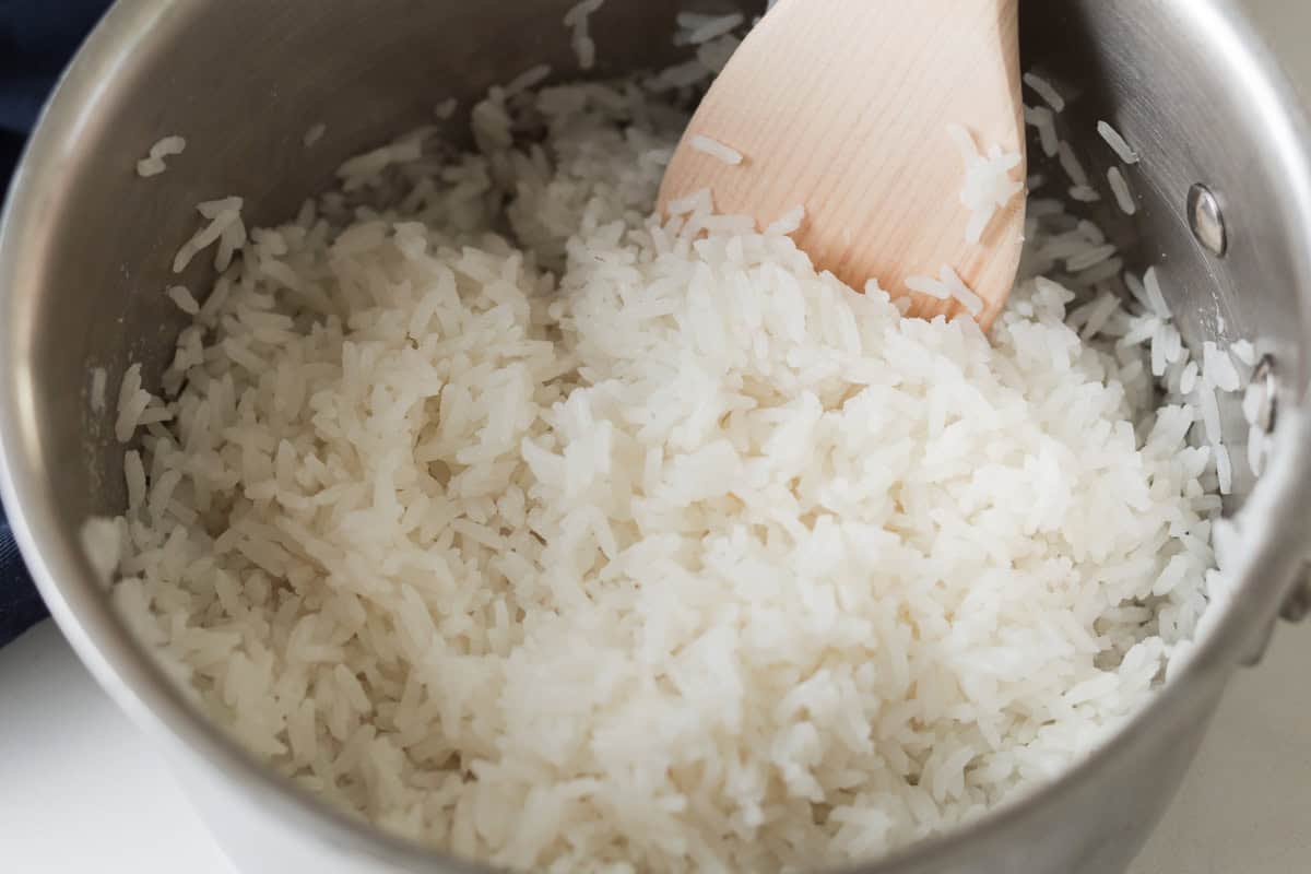 Freshly cooked, fluffy white jasmine rice inside a stainless steel pot with a wooden spoon fluffing the grains.