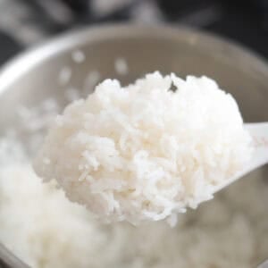 Close-up of a white rice paddle lifting a spoonful of fluffy, cooked white jasmine rice above a stainless steel pot.