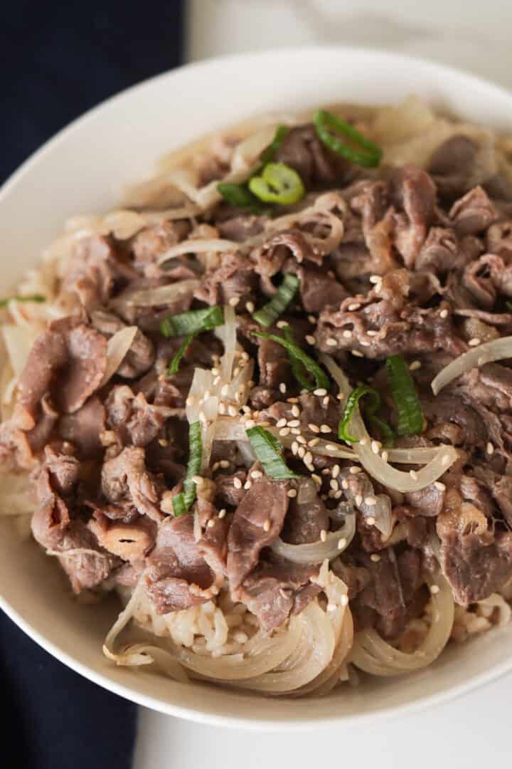 Vertical close-up shot of a white bowl filled with rice cooker Japanese beef bowl (gyudon), featuring tender thinly sliced beef, soft onions, sliced green onions, and toasted sesame seeds.