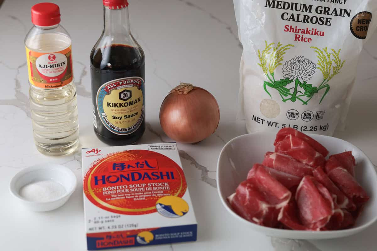 Flat-lay layout showing ingredients for rice cooker gyudon: Calrose rice, Kikkoman soy sauce, Aji-Mirin, HonDashi box, yellow onion, sugar, and thinly sliced beef.