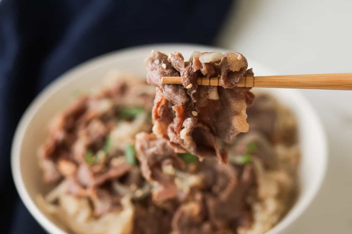Close-up shot of wooden chopsticks lifting a piece of tender sliced beef and onion from a bowl of Japanese gyudon.