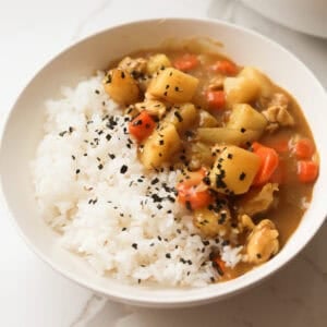 A white ceramic bowl filled with cooked Japanese chicken curry with cubed potatoes and carrots alongside white rice, sprinkled with black sesame seeds and furikake.