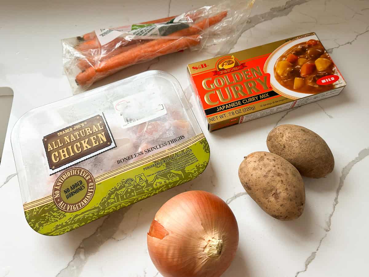 Flat-lay of raw ingredients for Japanese curry on a white countertop: a package of chicken thighs, carrots, whole yellow onion, two russet potatoes, and a box of S&B Golden Curry mix.