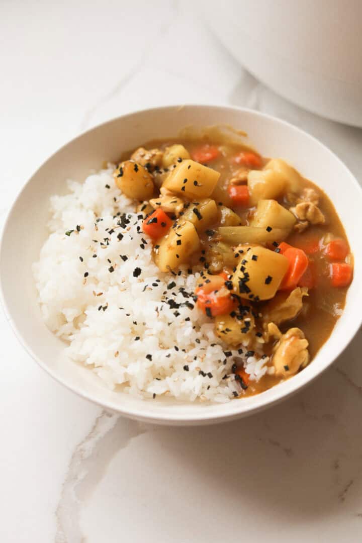 A served bowl of Japanese chicken curry over steamed white rice, garnished with furikake seasoning and black sesame seeds on a marble marble table.